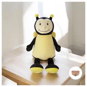 May include: A plush bee toy with black and yellow stripes, a smiling face, and yellow antennas. The bee is sitting on a wooden surface with a small heart-shaped box in the background.