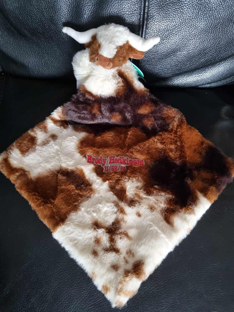 May include: A plush security blanket featuring a soft, brown and white cow head attached to a square, faux-fur blanket with a brown and white cow-print pattern. The blanket is embroidered with the red text Brody Hodkinson and the date 11/08/23.