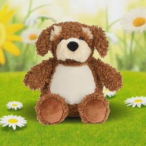 Può includere: Un giocattolo a forma di cane di peluche marrone con petto e muso bianchi, naso nero e orecchie flosce. L'animale di peluche è seduto su erba verde con margherite bianche e un fiore giallo sullo sfondo.