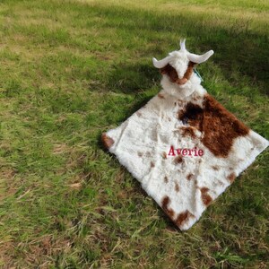May include: A plush security blanket featuring a stuffed cow head with white and brown fur. The square blanket body has a white and brown cow print pattern and the name Averie is embroidered in red thread in the center of the fabric.