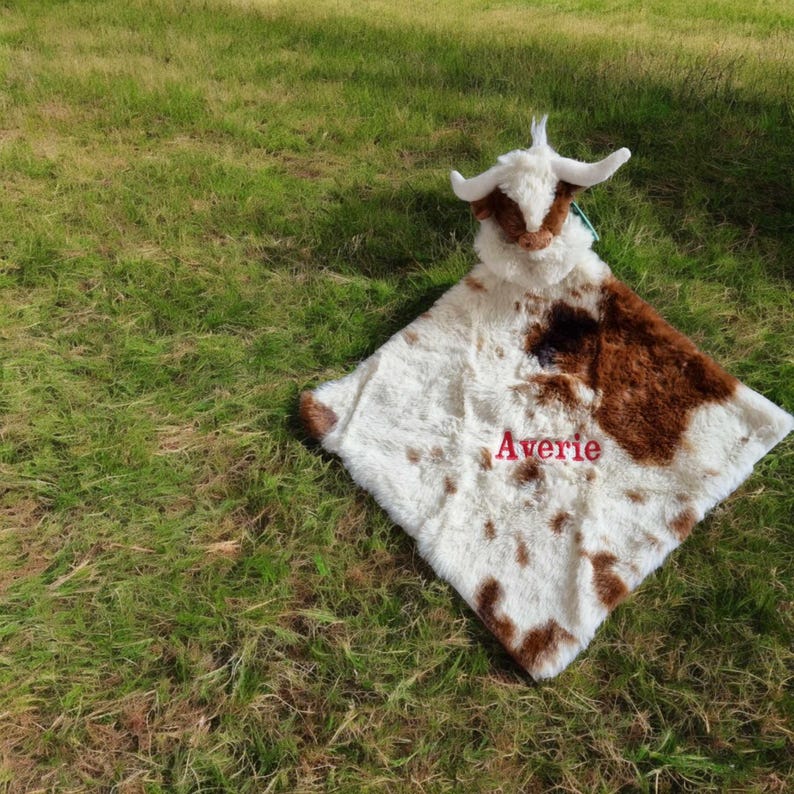 May include: A plush security blanket featuring a stuffed cow head with white and brown fur. The square blanket body has a white and brown cow print pattern and the name Averie is embroidered in red thread in the center of the fabric.