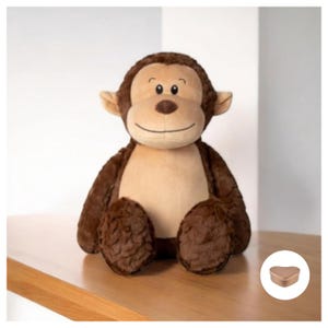 Op de afbeelding: Een bruin pluche aapje met een glimlachend gezicht en bruine vacht. De aap zit op een houten oppervlak met een klein hartvormig doosje in de hoek.