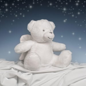 Könnte beinhalten: Ein weißer Teddybär mit Engelsflügeln sitzt auf weißem Stoff. Der Bär hat ein lächelndes Gesicht und befindet sich vor einem blauen Hintergrund mit Sternen. Ein weiches und kuscheliges Plüschtier.