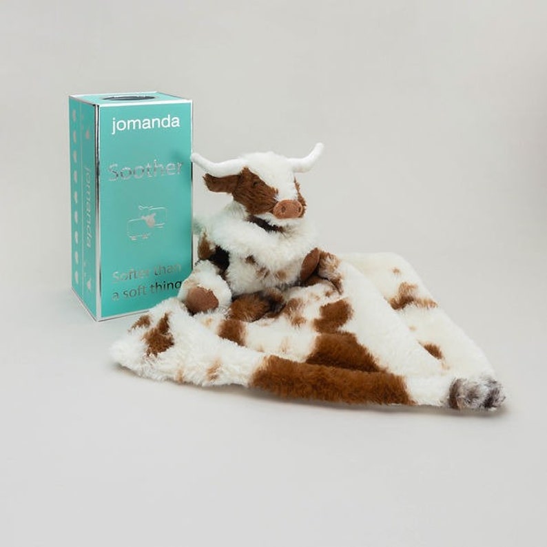 May include: A plush cow-themed security blanket with white and brown faux fur, featuring a stuffed cow head with horns. Beside it stands a rectangular turquoise box labeled with the text jomanda, Soother, and Softer than a soft thing.