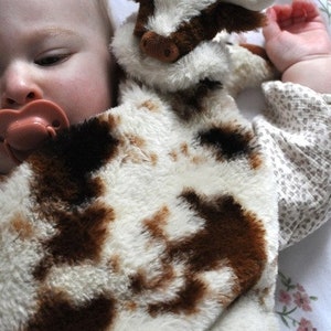 May include: A plush security blanket featuring a soft, fuzzy cow head with white horns and a brown and white spotted pattern. The fabric has a thick, textured pile, and the design includes a stuffed animal head attached to a flat, blanket-like body.