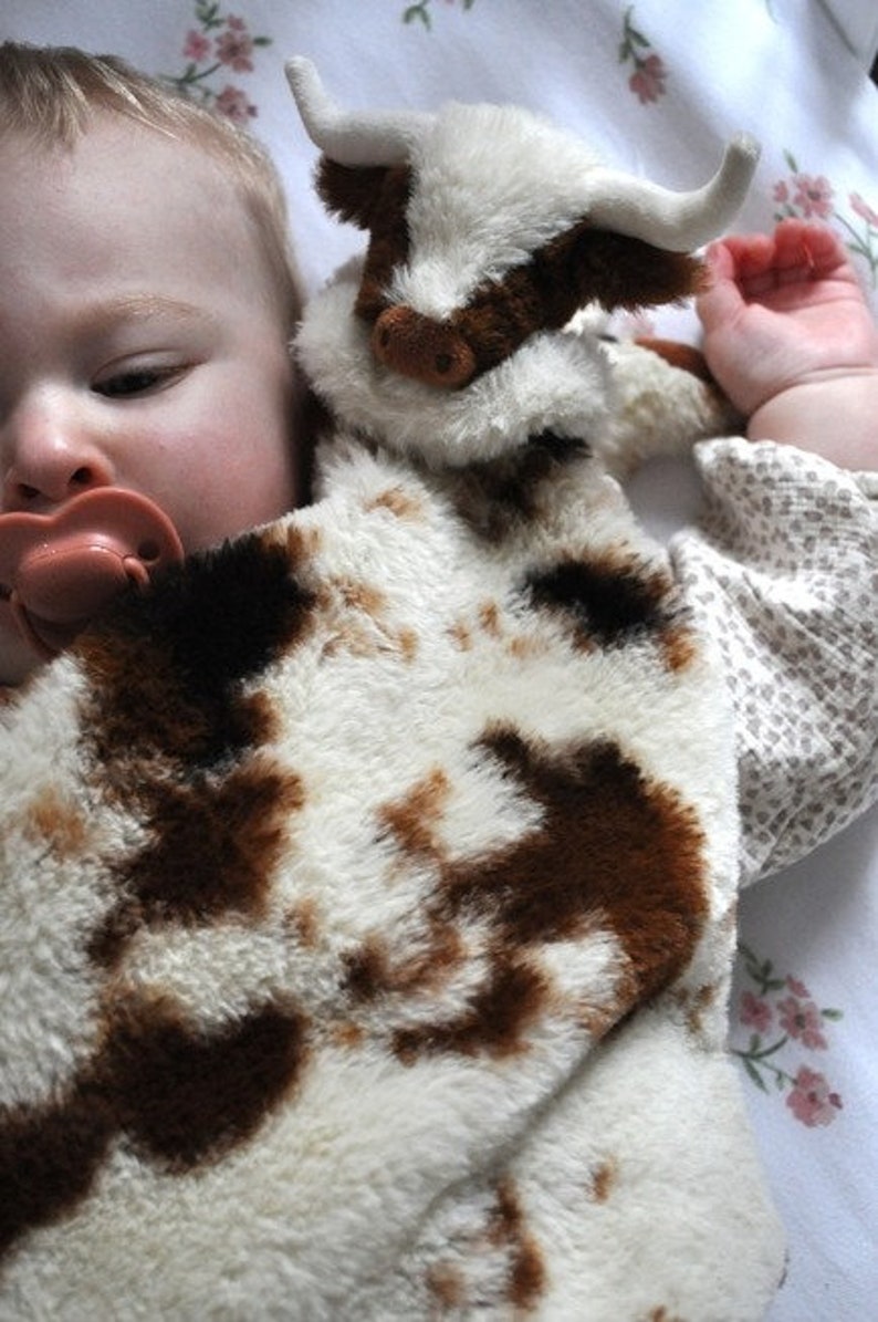 May include: A plush security blanket featuring a soft, fuzzy cow head with white horns and a brown and white spotted pattern. The fabric has a thick, textured pile, and the design includes a stuffed animal head attached to a flat, blanket-like body.