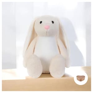 May include: A white plush bunny rabbit toy with a pink nose and black eyes. The bunny is sitting on a wooden surface. A small rose gold heart-shaped box is in the lower right corner of the image.