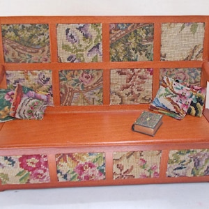 May include: A miniature wooden bench featuring a reddish-brown frame with inset panels of floral needlepoint fabric. The bench includes two small matching needlepoint cushions and a tiny, closed rectangular book resting on the seat surface.