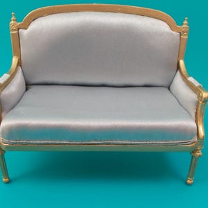 May include: A miniature sofa featuring a metallic gold frame with ornate detailing and smooth, light gray fabric upholstery on the seat, backrest, and armrests. The piece has four slender legs and a curved top edge on the backrest frame.