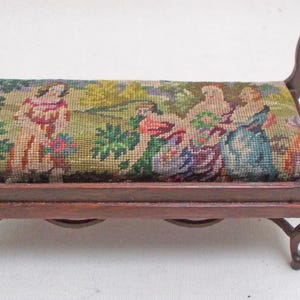 May include: A miniature wooden bench featuring a rectangular seat upholstered in a multi-colored needlepoint fabric depicting figures in a landscape. The bench has curved armrests and four small, ornate legs with a dark brown wood finish.