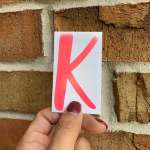May include: A bright pink letter K sticker on a white background. The sticker is being held by a hand.