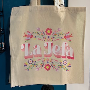 May include: A white canvas tote bag with a pink, white, and blue floral design and the text "La Jefa" in a pink and white gradient.