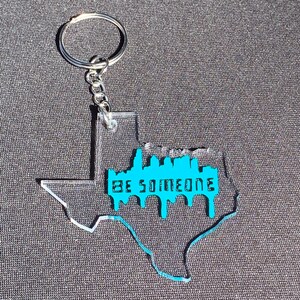 Könnte beinhalten: Ein klarer Acryl-Schlüsselanhänger in Texas-Form mit einem silbernen Schlüsselring. Der Anhänger hat ein türkisfarbenes Skyline-Design und die Worte "BE SOMEONE" in Türkis.