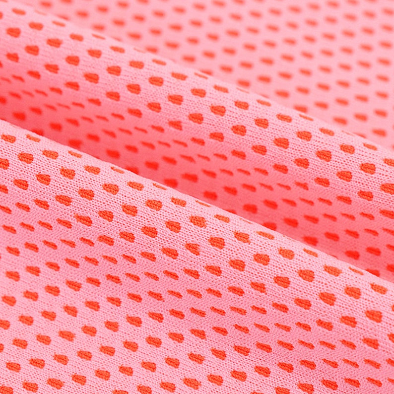May include: Close-up of a pink fabric with a dotted pattern in a darker shade of red. The fabric is folded, showing the texture and the arrangement of the dots. The material appears to be lightweight and breathable.