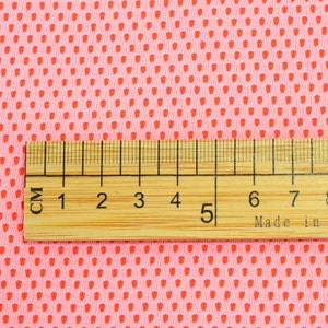 May include: Close-up of pink fabric with a repeating pattern of small, red, teardrop-shaped perforations. A wooden ruler with centimeter markings is placed across the fabric, showing measurements up to 8 cm.