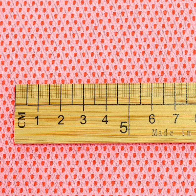 May include: Close-up of pink fabric with a repeating pattern of small, red, teardrop-shaped perforations. A wooden ruler with centimeter markings is placed across the fabric, showing measurements up to 8 cm.