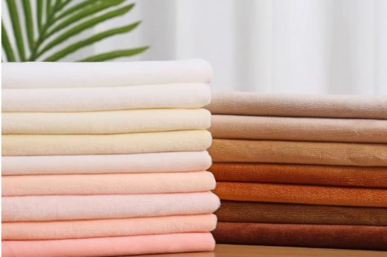 K&ouml;nnte beinhalten: Ein Stapel weicher, pl&uuml;schiger Stoffmuster in verschiedenen Beige-, Rosa- und Braunt&ouml;nen. Der Stoff ist ordentlich gefaltet und in einem Farbverlauf angeordnet.