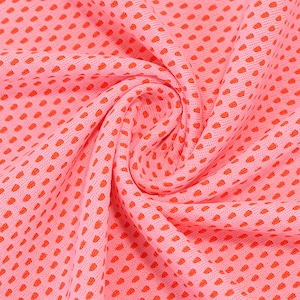May include: Close-up of a pink fabric with a repeating pattern of small, orange dots. The fabric is slightly gathered, creating soft folds and shadows. The texture appears to be breathable, possibly for athletic wear or summer clothing.