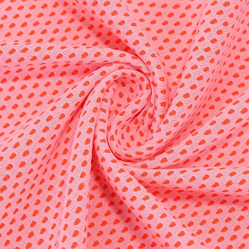 May include: Close-up of a pink fabric with a repeating pattern of small, orange dots. The fabric is slightly gathered, creating soft folds and shadows. The texture appears to be breathable, possibly for athletic wear or summer clothing.
