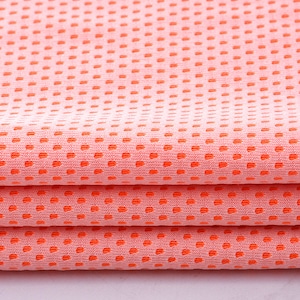 May include: Close-up of folded peach-colored fabric with a dotted pattern in a vibrant orange hue. The fabric is layered, showcasing the texture and design. The material appears to be lightweight and breathable, suitable for clothing or accessories.