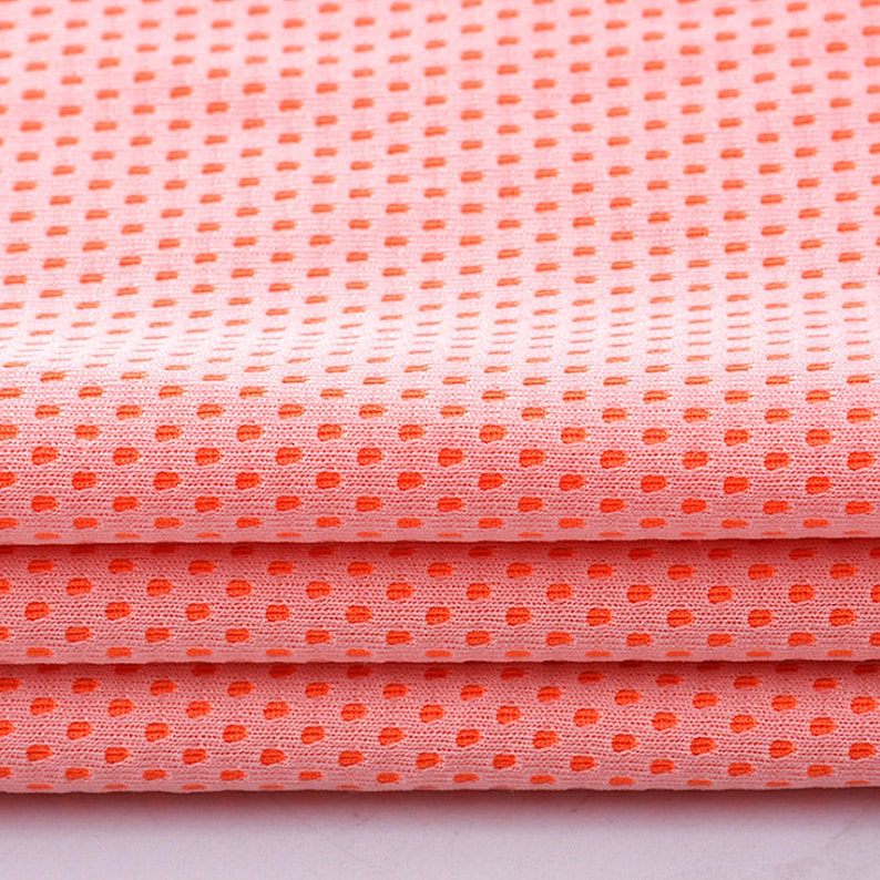 May include: Close-up of folded peach-colored fabric with a dotted pattern in a vibrant orange hue. The fabric is layered, showcasing the texture and design. The material appears to be lightweight and breathable, suitable for clothing or accessories.