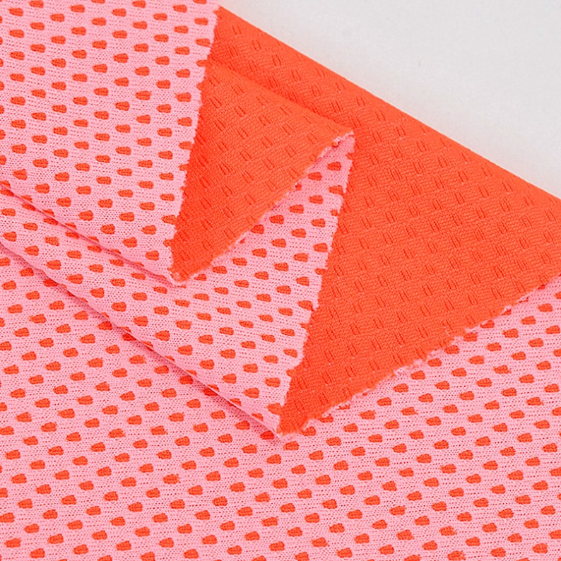 May include: Close-up of two pieces of fabric. One is a light pink with small, evenly spaced, red heart-shaped perforations. The other piece is a solid orange with a textured, grid-like pattern. The fabrics are layered, showcasing the contrast in color and texture.