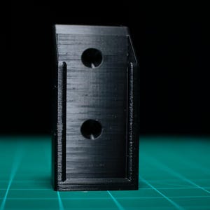 May include: A black plastic 3D printed object with two holes. The object is sitting on a green surface.