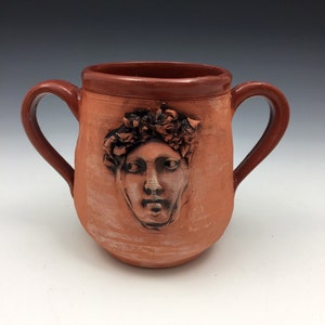 Könnte beinhalten: Eine Terrakotta-Tasse mit zwei Henkeln und einem Relief eines Gesichts mit einer Blumenkrone. Die Tasse ist rotbraun und das Gesicht hellbraun.