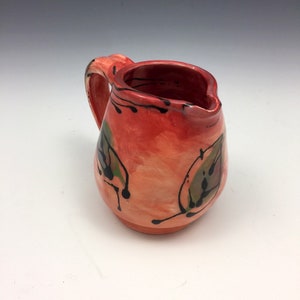 Pode incluir: Um pequeno jarro de cerâmica vermelho com uma alça. O jarro tem um design de folhas pretas e verdes.