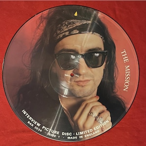 Peut inclure: Un disque vinyle transparent avec un portrait d'un homme portant des lunettes de soleil et un bandana. Le texte "THE MISSION" est imprimé à droite. Le disque est étiqueté "INTERVIEW PICTURE DISC - LIMITED EDITION" et "MADE IN ENGLAND".