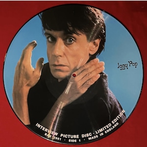 Könnte beinhalten: Eine runde Bildplatte mit Iggy Pop vor blauem Hintergrund. Das Bild zeigt Iggy Pop mit erhobener Hand, und der Text "Iggy Pop" befindet sich in der oberen rechten Ecke. Die Platte trägt den Text "INTERVIEW PICTURE DISC LIMITED EDITION MADE IN ENGLAND".