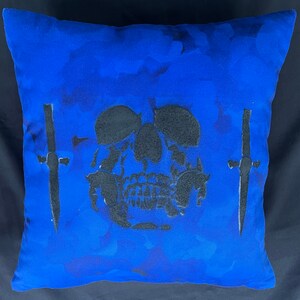 May include: Square blue decorative pillow with a black skull and two black daggers. The skull is centered on the pillow, and the daggers are on either side. The pillow has a dark blue background with lighter blue accents.