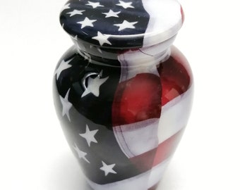 May include: A small, white ceramic urn with an American flag design. The urn has a lid and is decorated with red, white, and blue stripes and stars.