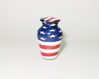 May include: A small, white ceramic urn with a red, white, and blue American flag design. The urn is about 4 inches tall and has a round shape.