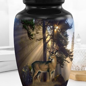 Puede incluir: Urna funeraria negra con una escena de bosque que presenta un ciervo en un claro soleado. La urna tiene un acabado brillante y una tapa segura. La obra de arte representa un sereno paisaje boscoso.