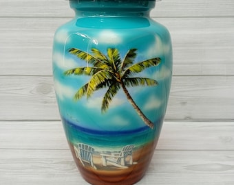 Könnte beinhalten: Eine tealblaue Keramikurne mit einer bemalten Szene eines Palmenbaums an einem Strand mit zwei weißen Stühlen. Die Urne hat einen Deckel.