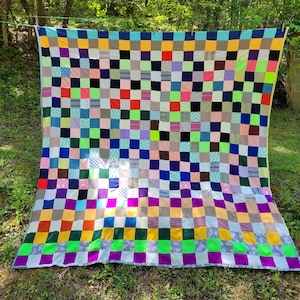 May include: A colorful patchwork quilt with a grid pattern of squares in various colors, including blue, green, purple, and yellow. The quilt is displayed outdoors, hanging on a clothesline, with a green grassy background.