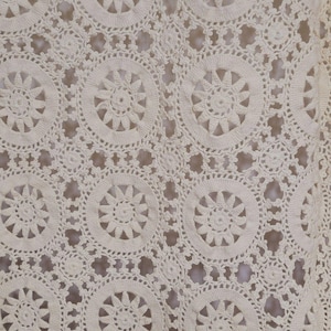 May include: An off-white crochet fabric featuring a repeating pattern of large circular motifs with sunburst centers, connected by smaller circular elements and openwork mesh, creating a continuous, intricate, and textured textile design.
