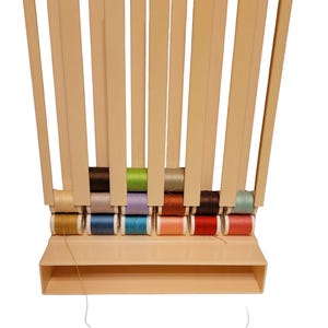 以下が含まれることがあります： ベージュのプラスチック製の糸立てに、金、青、緑、赤、ピンクなど、さまざまな色の糸巻きが12個あります。糸立ての下には小さな棚があります。