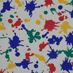 May include: Close-up of white fabric featuring a colourful paint splatter design. The splatters are red, blue, green, and yellow, creating a vibrant pattern. The fabric appears to be cotton or canvas.