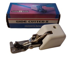 May include: A white and silver side cutter attachment for a sewing machine. The attachment is designed for zig-zag stitching. The box in the background is blue and red with the text "SIDE CUTTER II" and "Cutting & Sewing".
