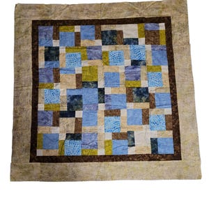 Może przedstawiać: Ręcznie robiona kołdra z patchworkowym wzorem. Kołdra posiada kwadraty z niebieskich, zielonych, brązowych i beżowych tkanin. Ciemnobrązowa obwódka obramowuje centralny wzór, a wzorzysta beżowa tkanina tworzy zewnętrzną obwódkę.