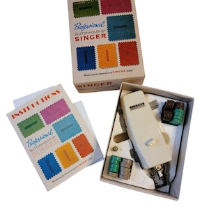 Puede incluir: Una máquina de coser Singer Professional Buttonholer vintage en su caja original. La caja es marrón con muestras de tela de colores y el texto "Professional Buttonholer by Singer". La máquina es blanca con un mango negro y un pedal de accionamiento plateado. La caja también incluye un conjunto de accesorios de plástico verde y marrón.