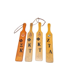 May include: Four wooden paddles with Greek letters and fraternity names. The paddles are brown and have black lettering. The letters on the paddles are Phi Sigma Kappa, Phi Kappa Tau, Phi Kappa Tau, and Zeta Tau Alpha.