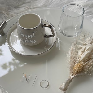 May include: White ceramic tray with a silver rim, featuring a white coffee cup with a silver handle and the text "Damat Kahvesi" in silver lettering. The tray also includes a clear glass, a small bouquet of dried white flowers, and the letters "N/O" in silver lettering.