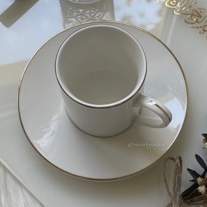 May include: A white ceramic coffee cup with a gold rim sits on a matching saucer. The cup and saucer are on a white surface.
