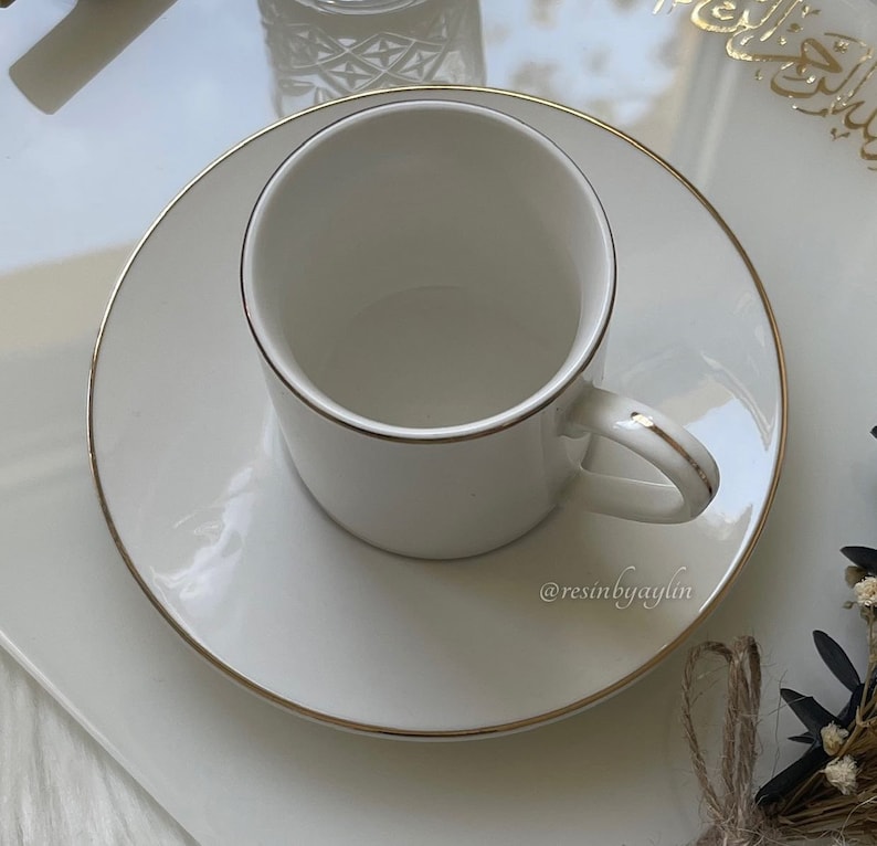 May include: A white ceramic coffee cup with a gold rim sits on a matching saucer. The cup and saucer are on a white surface.