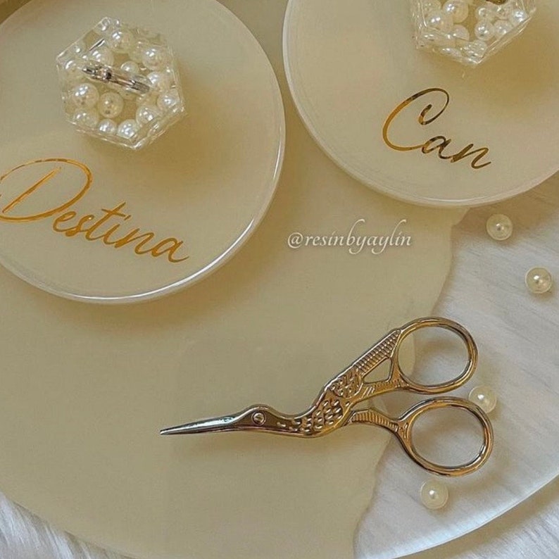 May include: Two white resin trays with gold lettering, one says "Destina" and the other says "Can". A small clear container with white pearls is on the "Destina" tray. A pair of silver scissors with a bird design is on the white surface.  The trays are on a white surface with white pearls scattered around.
