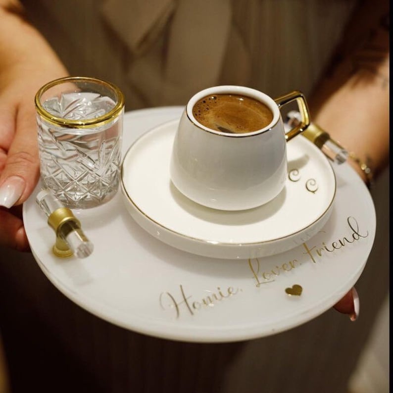 May include: White ceramic tray with gold trim and the words "Homie, Lover, Friend" written in gold script. The tray has a small heart design and holds a white ceramic cup with gold trim filled with coffee and a clear glass with water.