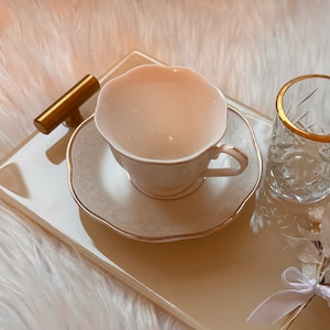 May include: A white porcelain teacup and saucer with a gold trim, sitting on a beige tray with a gold handle. The tray is on a white fluffy surface.